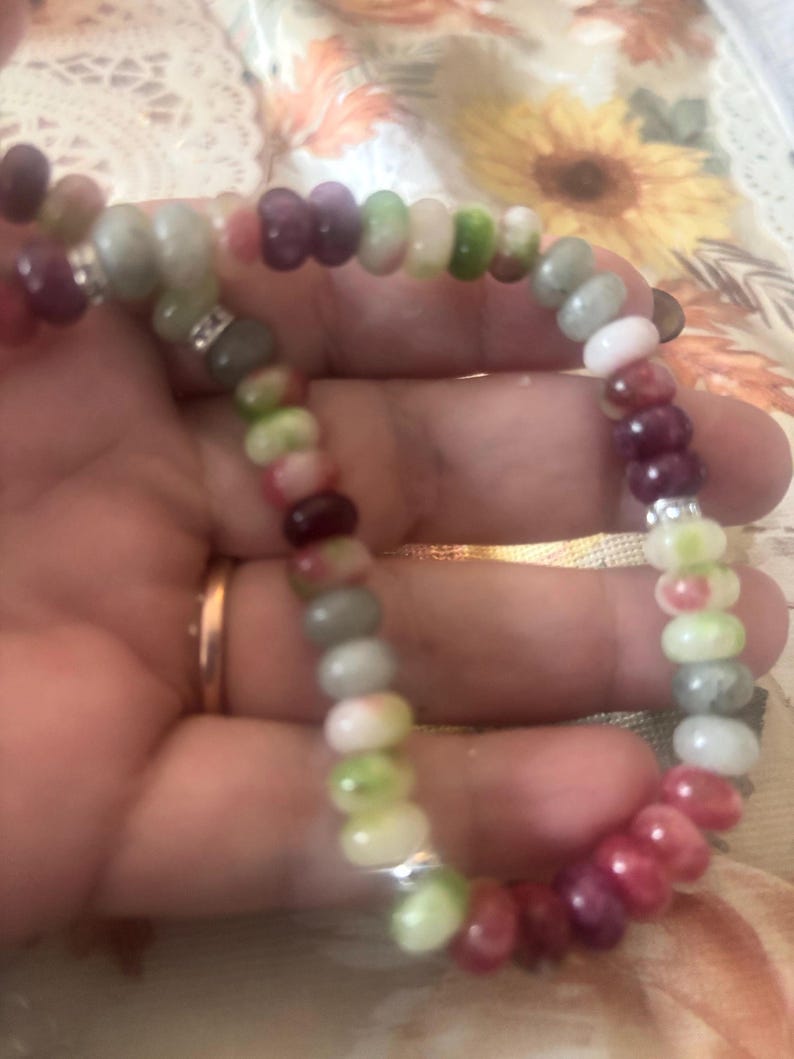 Puede incluir: Una pulsera de cuentas coloridas con cuentas de piedras preciosas en tonos rojos, rosas, verdes y grises. La pulsera est&aacute; ensartada en un cord&oacute;n flexible e incluye detalles plateados. Las cuentas miden aproximadamente 0,6 cm de di&aacute;metro.