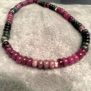 Peut inclure: Un collier de perles compos&eacute; de perles roses fonc&eacute;es, vertes et noires. Des entretoises argent&eacute;es s&eacute;parent les perles. Le collier est pos&eacute; sur une surface grise. Les perles sont probablement des pierres pr&eacute;cieuses.