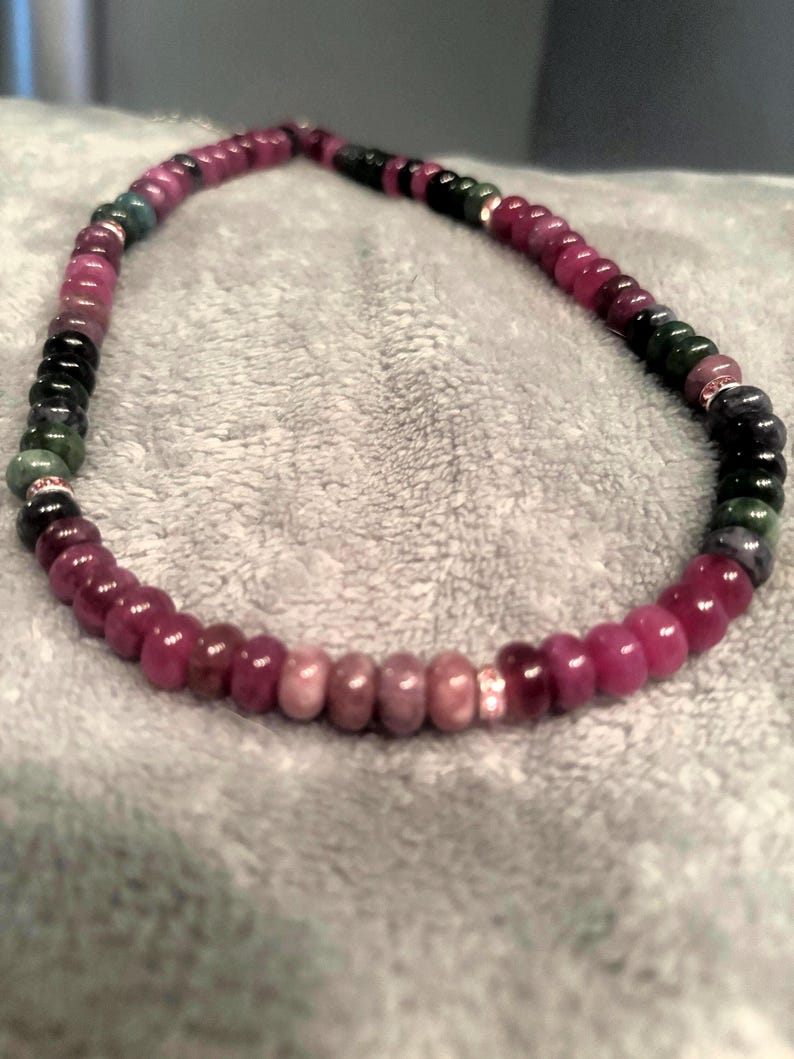 Peut inclure: Un collier de perles compos&eacute; de perles roses fonc&eacute;es, vertes et noires. Des entretoises argent&eacute;es s&eacute;parent les perles. Le collier est pos&eacute; sur une surface grise. Les perles sont probablement des pierres pr&eacute;cieuses.