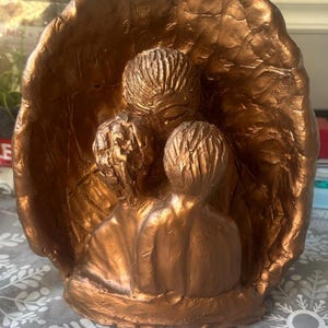 Puede incluir: Una escultura de color bronce que representa a una familia de tres figuras. Las figuras están agrupadas, con la figura central que parece abrazar a las otras dos. La escultura tiene una superficie texturizada y está colocada sobre un fondo que se asemeja a una gran hoja estilizada.