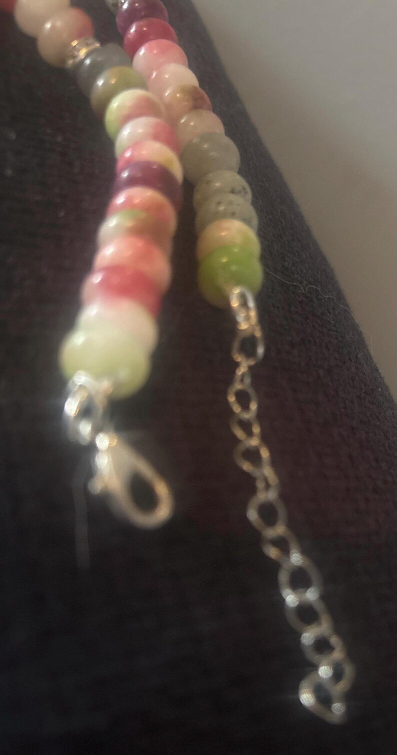 Puede incluir: Primer plano de un collar de cuentas de colores. El collar presenta una variedad de cuentas redondas en tonos rosa, verde y blanco. El collar tiene una cadena y un cierre de color plateado.