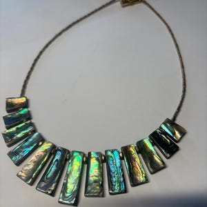 Può includere: Collana con pendenti rettangolari iridescenti in conchiglia di abalone nei toni del verde, blu e oro. I pendenti sono infilati su una delicata catena argentata con chiusura dorata. La collana è esposta su una superficie bianca.
