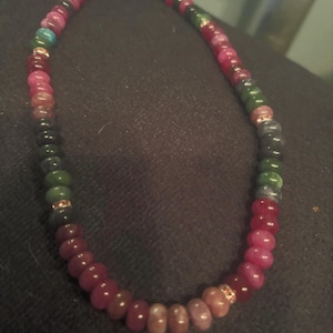 Peut inclure: Un collier de perles avec une vari&eacute;t&eacute; de pierres pr&eacute;cieuses color&eacute;es. Le collier pr&eacute;sente un m&eacute;lange de perles rouges, roses, vertes et bleues, avec des accents dor&eacute;s. Les perles sont enfil&eacute;es pour cr&eacute;er un collier.