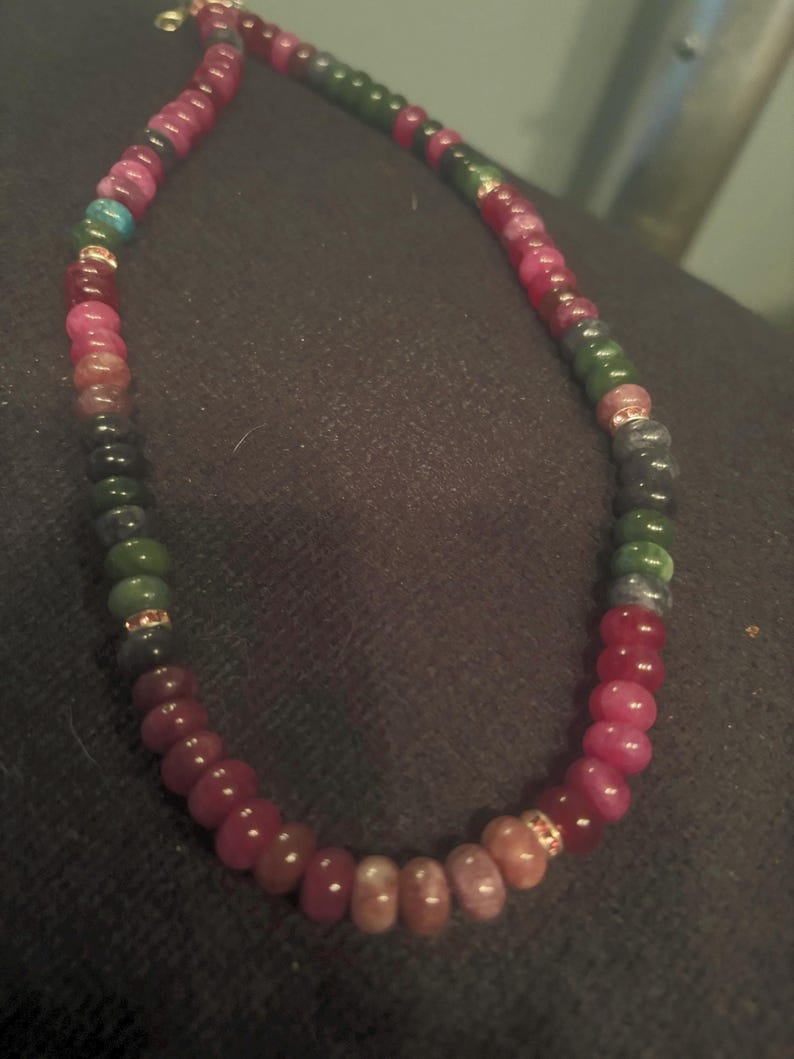 Peut inclure: Un collier de perles avec une vari&eacute;t&eacute; de pierres pr&eacute;cieuses color&eacute;es. Le collier pr&eacute;sente un m&eacute;lange de perles rouges, roses, vertes et bleues, avec des accents dor&eacute;s. Les perles sont enfil&eacute;es pour cr&eacute;er un collier.