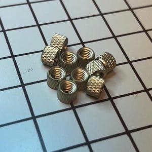 Puede incluir: Un conjunto de pequeñas tuercas moleteadas de color latón. Las tuercas tienen forma cilíndrica con rosca interna y un exterior texturizado para facilitar el agarre. Están dispersas sobre una superficie cuadriculada.