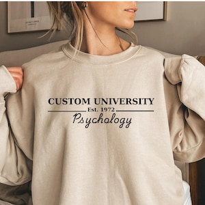 Könnte beinhalten: Beiges Sweatshirt mit dem schwarzen Text "CUSTOM UNIVERSITY Est. 1972 Psychology". Das langärmlige Sweatshirt hat eine neutrale Farbe. Der Text ist mittig auf der Vorderseite platziert.