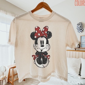 May include: Ivory-colored t-shirt featuring a graphic of Minnie Mouse with a red polka-dot bow. The Comfort Colors brand shirt is hanging on a wooden hanger. The shirt is a relaxed fit and has short sleeves.