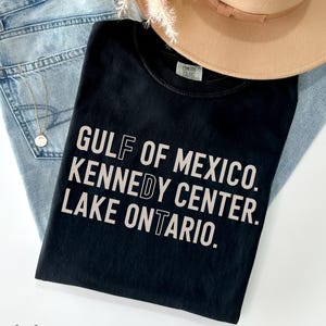 May include: A black t-shirt featuring the text GULF OF MEXICO. KENNEDY CENTER. LAKE ONTARIO. in light beige lettering, with specific letters F, D, and T highlighted in a white outline font, layered over light blue denim jeans and a tan hat.