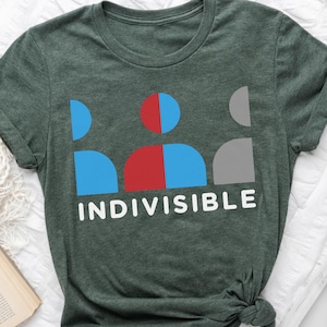May include: A dark green t-shirt with the word "INDIVISIBLE" in white block letters. Above the word is a graphic of a person made of blue, red, and gray shapes. The shirt is folded at the bottom.