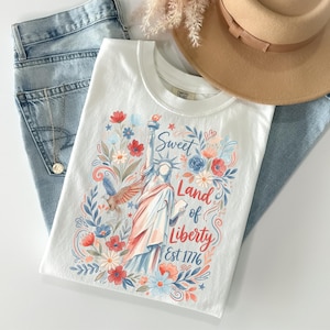 May include: White t-shirt featuring a colorful illustration of the Statue of Liberty, an eagle, and floral elements. The text reads "Sweet Land of Liberty Est 1776." The shirt is paired with blue jeans and a tan hat.