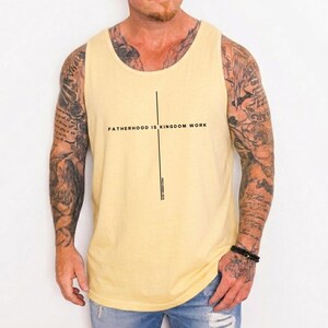 May include: A light yellow tank top with the text "FATHERHOOD IS KINGDOM WORK" in black, with a vertical line and the text "PROVERBS 20:7". The tank top is worn by a person with tattoos and blue jeans.
