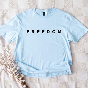 Op de afbeelding: Een lichtblauw t-shirt met het woord "FREEDOM" in zwarte blokletters gedrukt. Het shirt met korte mouwen is gemaakt van een zachte stof. Het shirt ligt op een wit en beige patroon.