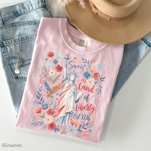 May include: A light pink t-shirt with a graphic of the Statue of Liberty, flowers, and the text "Sweet Land of Liberty Est 1776." The shirt is paired with blue jeans and a tan hat. A gold necklace is also visible.