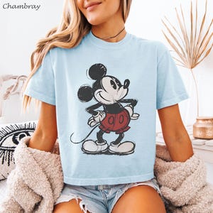 May include: Light blue t-shirt featuring a distressed graphic of Mickey Mouse. The cartoon character is depicted in black, white, and red. The shirt has short sleeves and a crew neck. The word "Chambray" is in the top left corner.