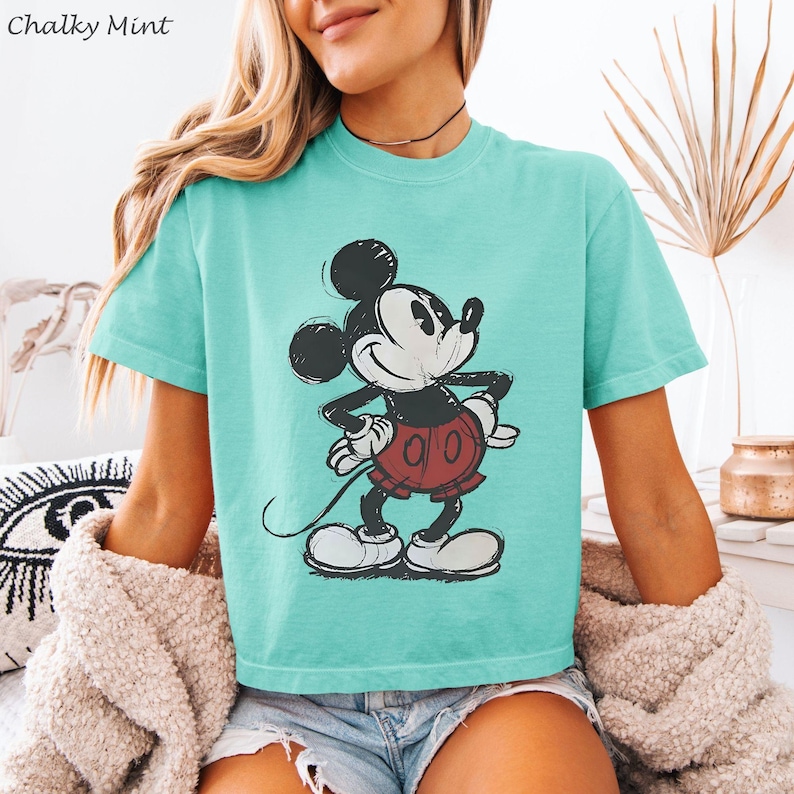 May include: A turquoise t-shirt featuring a distressed graphic of Mickey Mouse. The cartoon character is depicted in black, white, and red. The text "Chalky Mint" is in the upper left corner. The shirt has short sleeves.