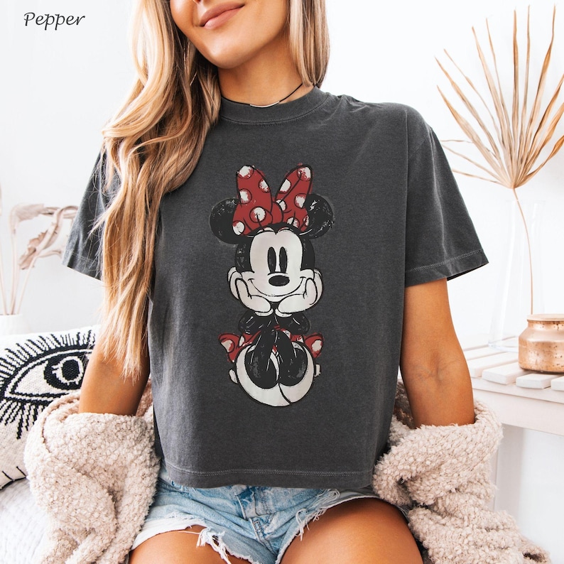 May include: Dark gray t-shirt featuring a distressed Minnie Mouse graphic with a red polka dot bow. The shirt has a crew neck and short sleeves. The word "Pepper" is in the top left corner.