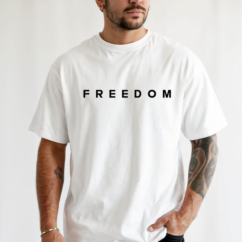 Op de afbeelding: Wit T-shirt met het woord "FREEDOM" in zwart gedrukt op de borst. Het shirt heeft korte mouwen en een ronde hals. Het model draagt het shirt.
