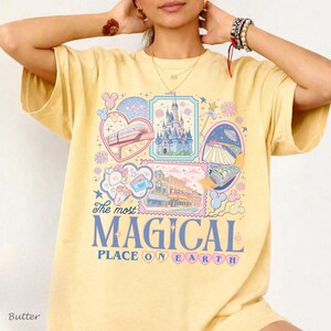 Pu&ograve; includere: T-shirt giallo burro con un design grafico colorato. Il design include illustrazioni di un castello, una monorotaia e altri elementi del parco a tema, con la scritta "The most MAGICAL PLACE ON EARTH".