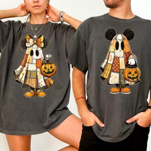 May include: Two dark gray t-shirts featuring a cartoon ghost character with Mickey Mouse ears, a patchwork design, and a jack-o'-lantern. The ghost is wearing orange shoes and holding a pumpkin bucket. The shirts are oversized.