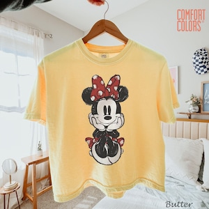 May include: Butter yellow t-shirt featuring a graphic of Minnie Mouse with a red polka dot bow. The shirt is hanging on a wooden hanger. The text "COMFORT COLORS" is visible in the upper right corner.