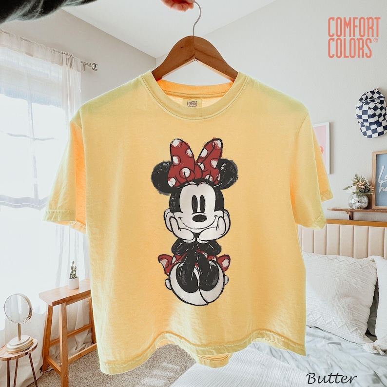 May include: Butter yellow t-shirt featuring a graphic of Minnie Mouse with a red polka dot bow. The shirt is hanging on a wooden hanger. The text "COMFORT COLORS" is visible in the upper right corner.
