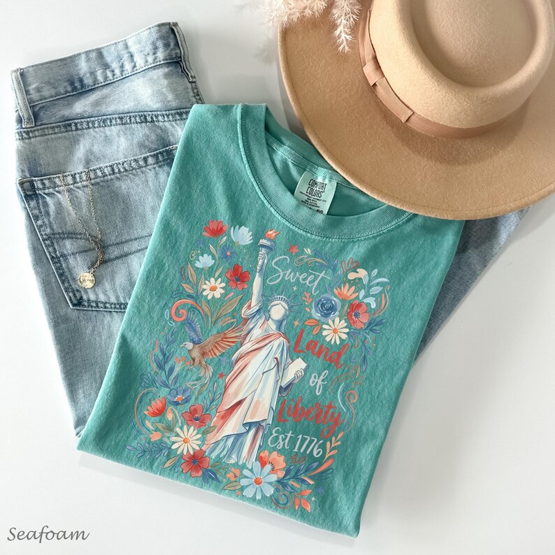 May include: A seafoam green t-shirt with a graphic of the Statue of Liberty, surrounded by flowers and the text "Sweet Land of Liberty Est 1776." The shirt is paired with light wash jeans, a gold necklace, and a tan fedora hat.