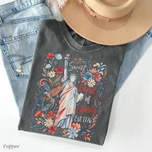 May include: A charcoal gray t-shirt featuring a colorful graphic of the Statue of Liberty surrounded by flowers and the text "Sweet Land of Liberty Est 1776." The shirt is paired with light blue jeans and a tan hat.