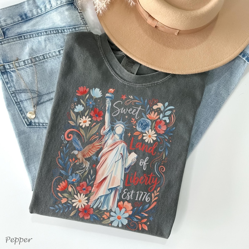 May include: A charcoal gray t-shirt featuring a colorful graphic of the Statue of Liberty surrounded by flowers and the text "Sweet Land of Liberty Est 1776." The shirt is paired with light blue jeans and a tan hat.