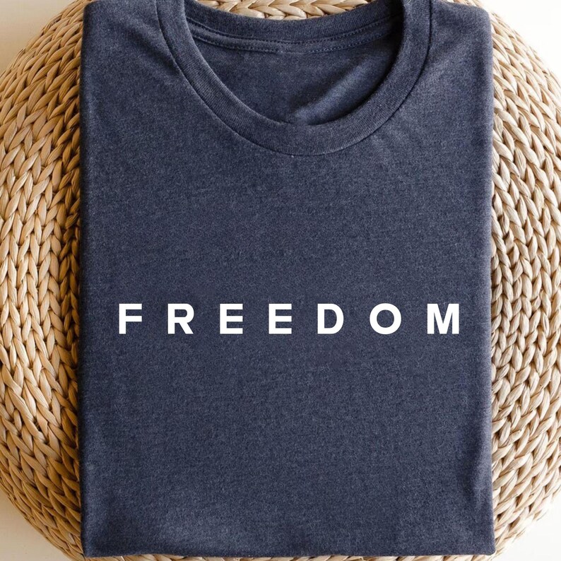 Op de afbeelding: Een opgevouwen marineblauw t-shirt met het woord "FREEDOM" in witte blokletters. Het shirt ligt op een geweven, lichtbruin oppervlak. Het t-shirt heeft een ronde hals.