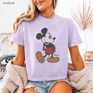 May include: A light purple t-shirt featuring a graphic of Mickey Mouse in a walking pose. Mickey Mouse is depicted in his classic attire: red shorts, brown shoes, and black gloves. The Disney logo is visible. The word "Orchid" is in the top left corner.