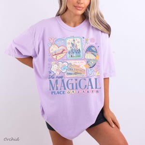 Pu&ograve; includere: Una maglietta lilla chiaro con un design grafico che presenta varie illustrazioni, tra cui un castello, un treno e altre immagini a tema. Il testo "The most MAGICAL PLACE ON EARTH" &egrave; visualizzato in blu e bianco. La parola "Orchid" &egrave; stampata in basso a sinistra.