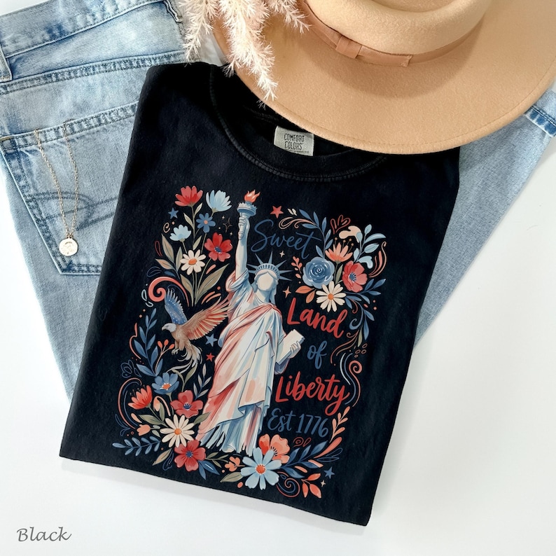 May include: Black t-shirt featuring a colorful graphic of the Statue of Liberty, flowers, and an eagle. The text reads "Sweet Land of Liberty Est 1776". A tan hat and blue jeans are in the background.