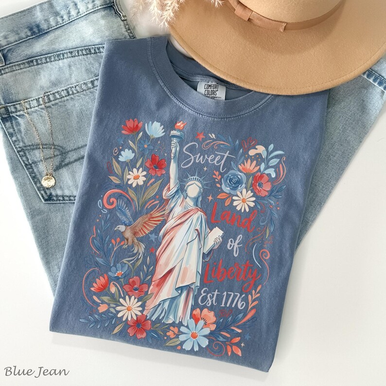May include: A blue t-shirt with a graphic of the Statue of Liberty, flowers, and the text "Sweet Land of Liberty Est 1776." The shirt is paired with blue jeans and a tan hat. A gold necklace is also visible.
