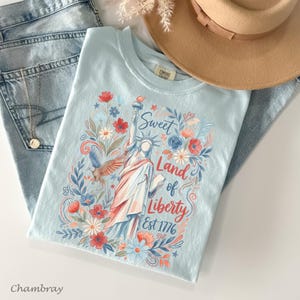 May include: A light blue t-shirt featuring a floral design with the Statue of Liberty, the text "Sweet Land of Liberty Est 1776", and a flying eagle. The shirt is paired with blue jeans and a tan hat.