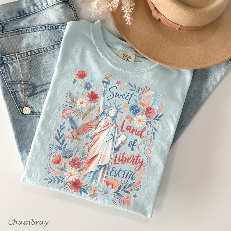 May include: A light blue t-shirt featuring a floral design with the Statue of Liberty, the text "Sweet Land of Liberty Est 1776", and a flying eagle. The shirt is paired with blue jeans and a tan hat.