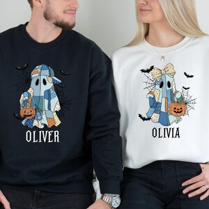 May include: Matching Halloween sweatshirts. One is navy blue, the other is white. Each features a patchwork ghost design holding a pumpkin and surrounded by bats and a spiderweb. The name "OLIVER" is printed on the navy sweatshirt, and "OLIVIA" on the white.
