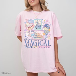 Pu&ograve; includere: T-shirt rosa chiaro con un design grafico colorato. Il design include illustrazioni di un castello, un treno e altri elementi stravaganti, insieme al testo "The most MAGICAL PLACE ON EARTH."