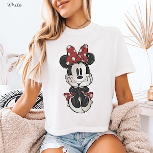 May include: White t-shirt featuring a distressed graphic of Minnie Mouse. Minnie is depicted with a red polka dot bow, black ears, and a red dress. The word "White" is printed in the upper left corner.