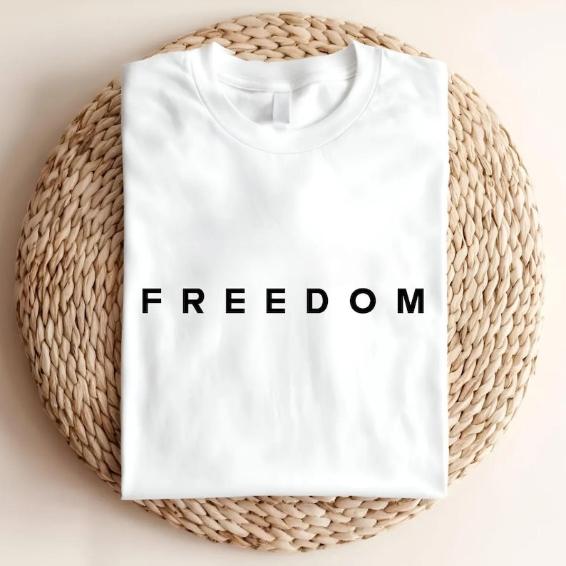 Op de afbeelding: Een wit t-shirt met het woord "FREEDOM" in zwarte letters in het midden. Het shirt is opgevouwen en ligt op een ronde, geweven placemat. De afbeelding heeft een strakke, minimalistische uitstraling.