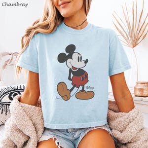 May include: Light blue t-shirt featuring a classic Mickey Mouse graphic. The cartoon character is depicted in his iconic red shorts, brown shoes, and black ears. The Disney logo is visible. The shirt is made of soft material.