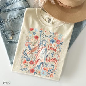 May include: Ivory-colored t-shirt featuring a colorful illustration of the Statue of Liberty, surrounded by flowers and the text "Sweet Land of Liberty Est 1776." The shirt is paired with light-wash denim jeans and a tan hat.