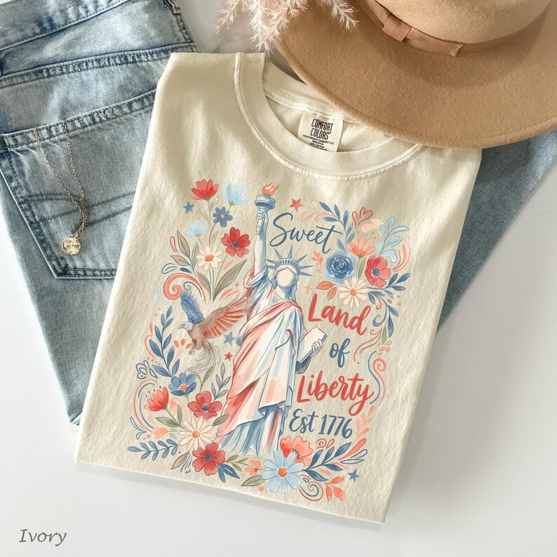 May include: Ivory-colored t-shirt featuring a colorful illustration of the Statue of Liberty, surrounded by flowers and the text "Sweet Land of Liberty Est 1776." The shirt is paired with light-wash denim jeans and a tan hat.