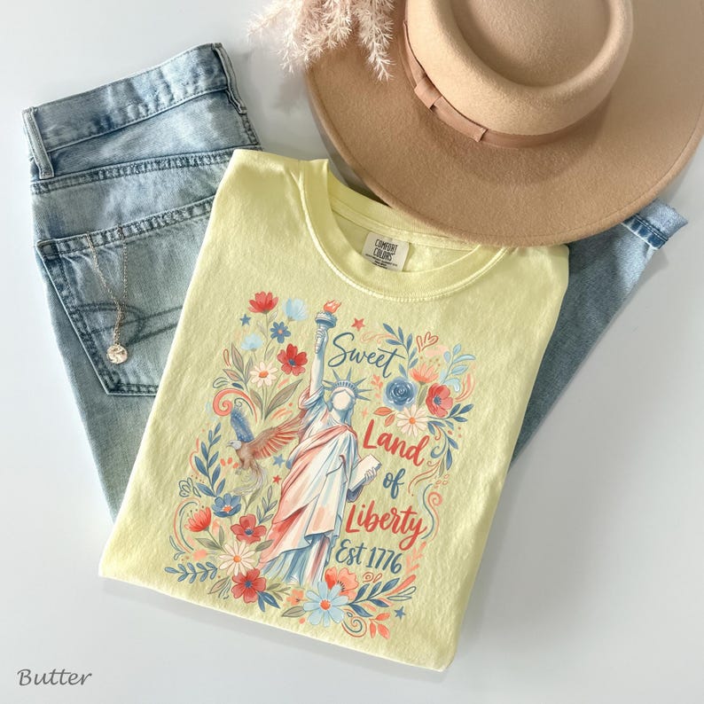 May include: A light yellow t-shirt with a graphic of the Statue of Liberty, surrounded by flowers and the text "Sweet Land of Liberty Est 1776." The shirt is paired with light blue jeans, a tan hat, and a necklace.
