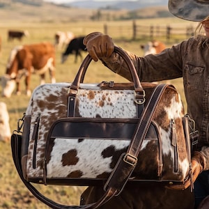 May include: A duffel bag crafted from brown and white cowhide leather featuring a large front zippered pocket, two top handles, and a detachable shoulder strap with antique brass hardware accents and dark brown leather trim details.