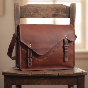 Op de afbeelding: Een bruine leren messenger bag met een klepsluiting, messing hardware en een verstelbare schouderriem. De tas staat op een houten stoel. De tas heeft een klassieke, vintage stijl.