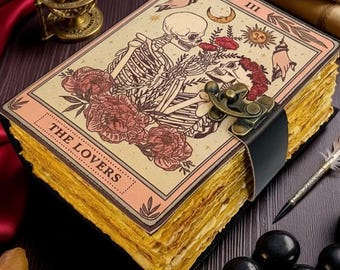 Op de afbeelding: Een vintage journal met een tarotkaartontwerp met skeletten en rozen. De omslag leest "The Lovers" en is vastgezet met een metalen sluiting en een zwarte leren band. De verouderde pagina's zijn zichtbaar, samen met een veer, een inktpot en decoratieve items.