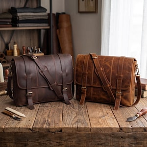 Puede incluir: Dos bolsos mensajeros rectangulares de cuero con correas ajustables y cierres de doble hebilla. Un bolso es de color marrón oscuro y el otro es de color marrón claro. Ambos presentan paneles cosidos verticales y detalles metálicos.