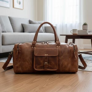 May include: A brown leather duffel bag featuring two top handles, a detachable shoulder strap, and a central front pocket with a buckle closure. The bag includes side zippered compartments and antique brass-toned metal hardware accents.