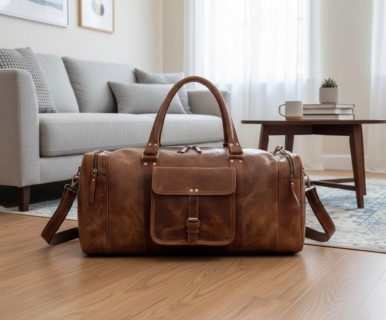 May include: A brown leather duffel bag featuring two top handles, a detachable shoulder strap, and a central front pocket with a buckle closure. The bag includes side zippered compartments and antique brass-toned metal hardware accents.