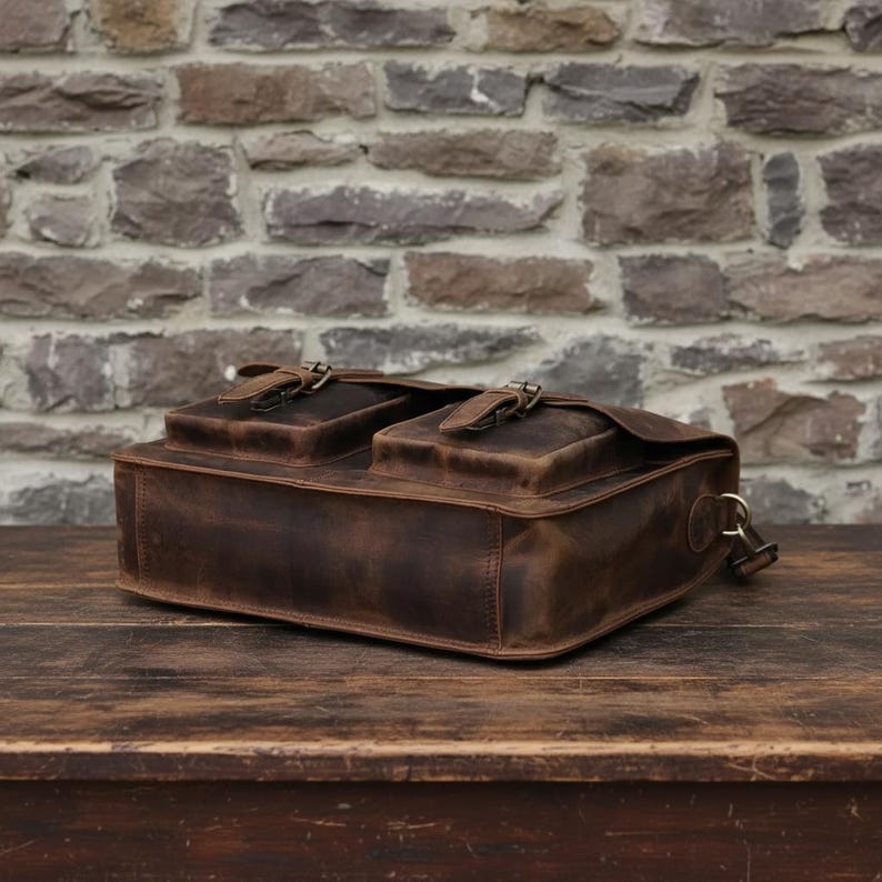 May include: A dark brown leather messenger bag featuring two front pockets with flap closures secured by metal buckles. The bag has a rectangular structure with visible stitching along the edges and a metal D-ring attachment on the side.